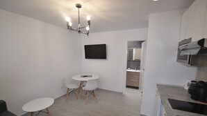 A modern kitchen with white cabinets, a built-in oven, and a coffee machine. There is a dining area with a small table and two chairs.