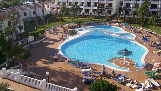 A resort with a central swimming pool surrounded by sun loungers and palm trees.
