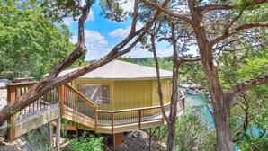 A wooden deck with a railing, a yellow building with a window, and a view of a lake surrounded by trees. - The Treehouse on Lake Travis (Lakeway)