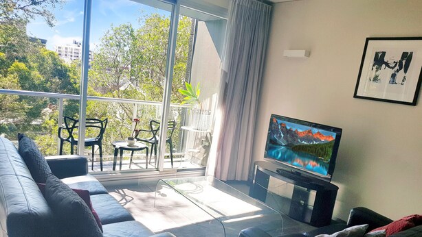 Sala de estar moderna com sofá, mesa de centro e televisão sobre um móvel. Há uma varanda com vista para árvores e edifícios.