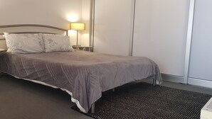 Cama bem arrumada com colcha cinza e duas almofadas, com duas luminárias de cabeceira em mesas de apoio brancas.