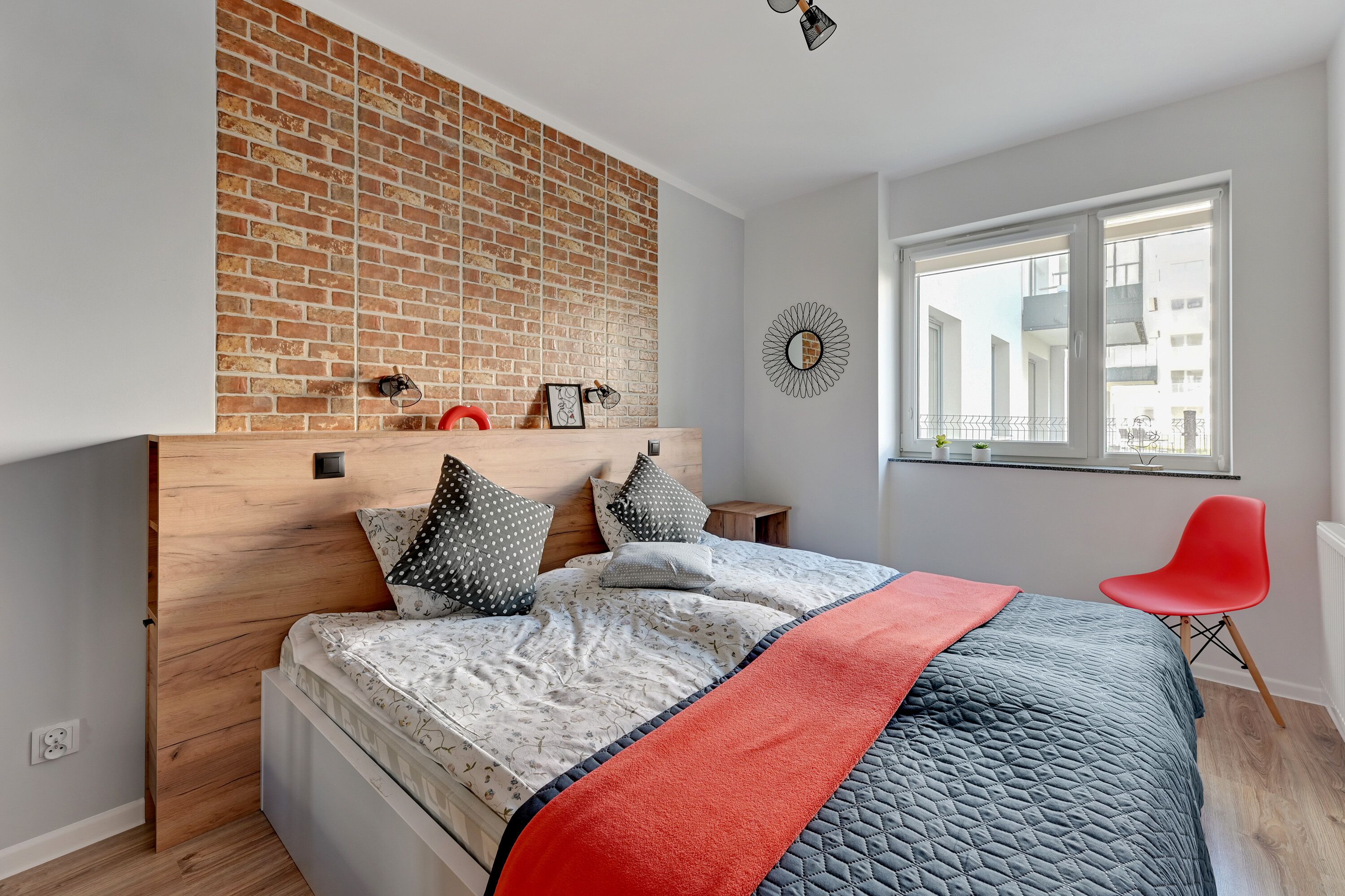 A modern bedroom with a brick accent wall, a wooden headboard, a large bed with patterned bedding, a red chair, and a window with a view of buildings.