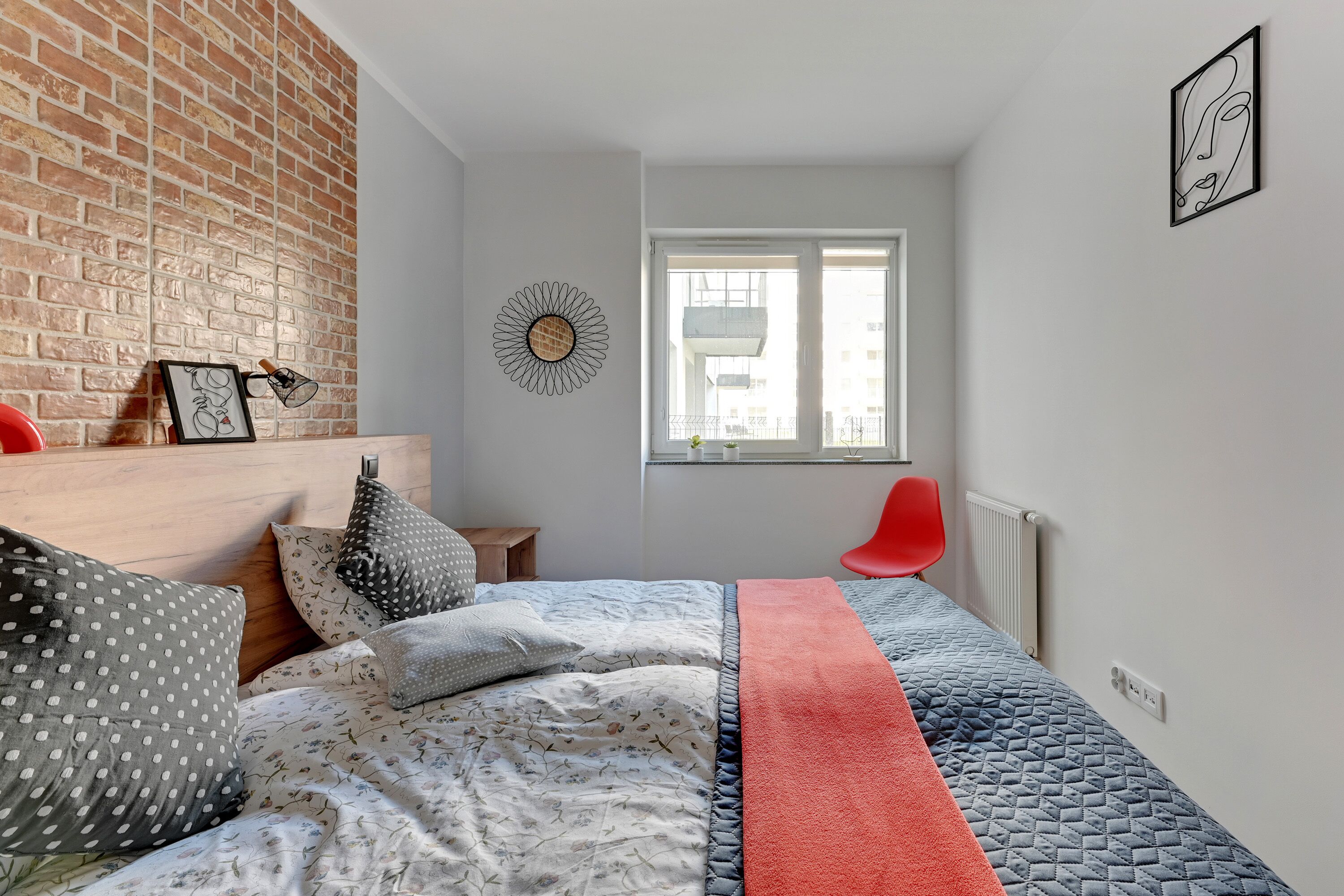 A bedroom with a brick wall, a bed with patterned bedding, a red chair, and a window with a view of buildings.