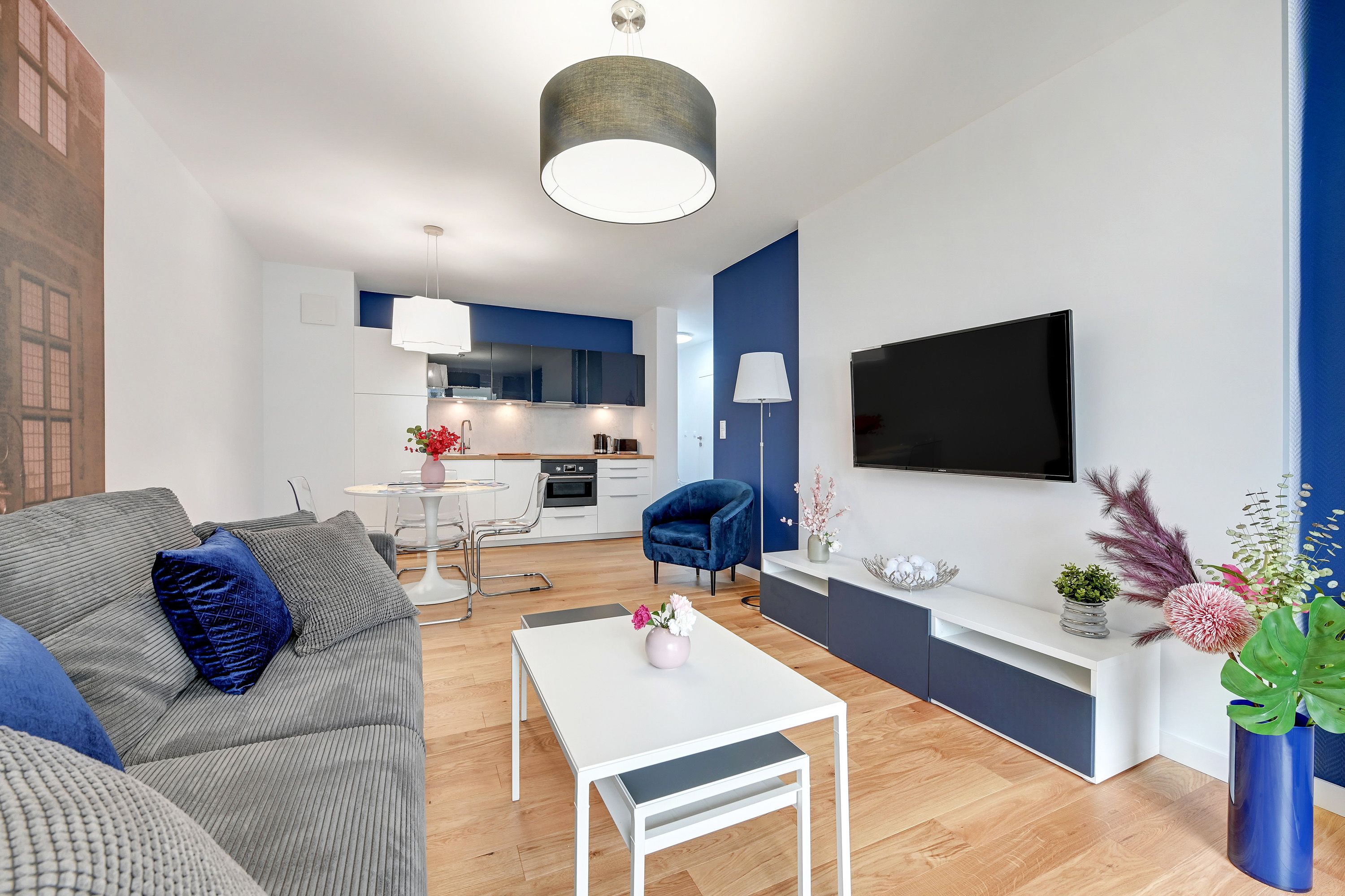 Un soggiorno moderno con un divano grigio, un tavolino da caffè bianco e una poltrona blu. Si intravede una cucina sullo sfondo, con una TV montata a parete.