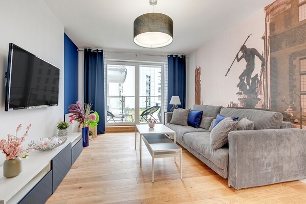 A modern living room with a flat-screen TV, a sofa, a coffee table, and a large window with curtains. - NORDBYHUS Apartamenty Angielska Grobla (Gdansk)