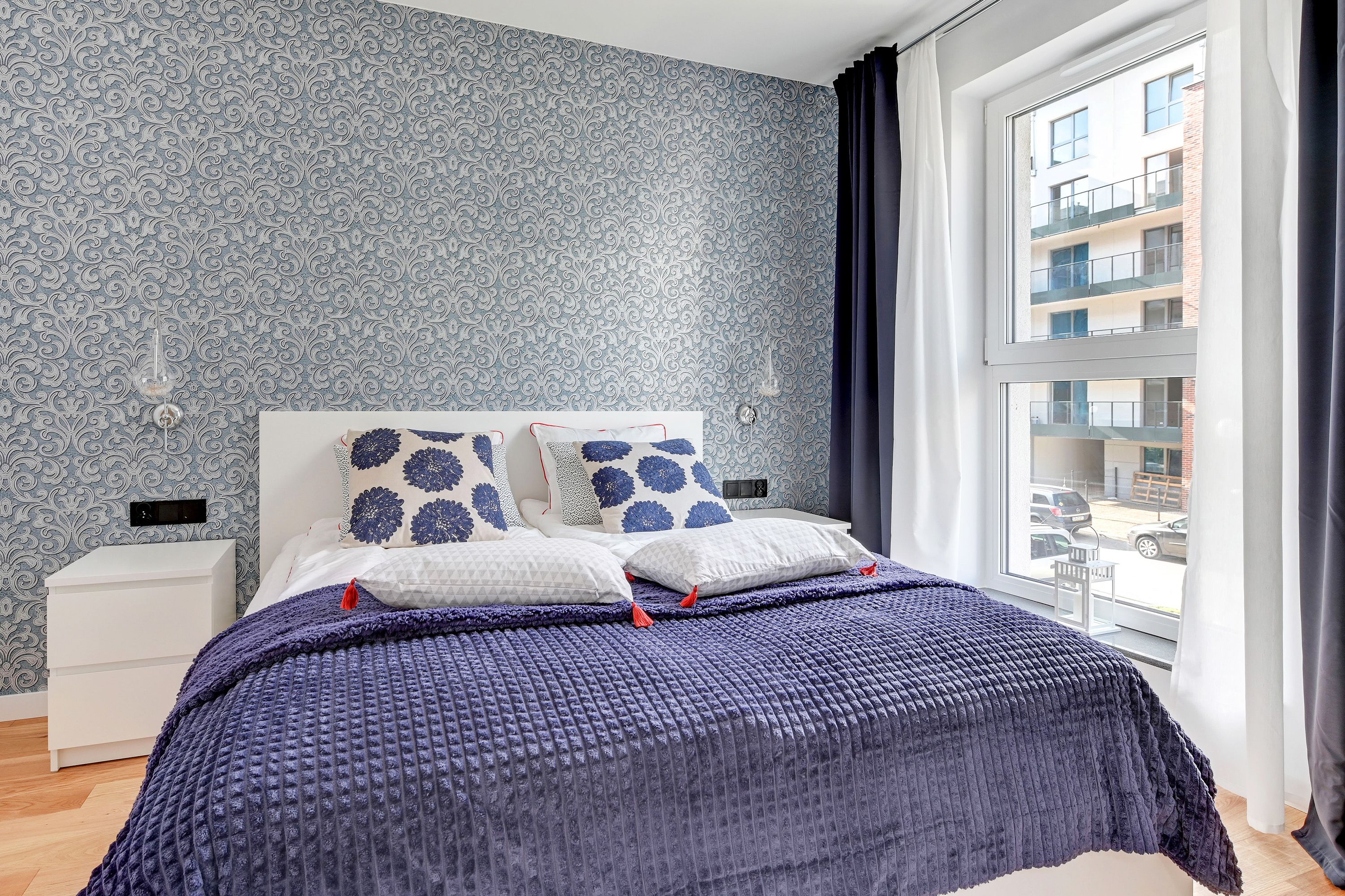 Une chambre avec un papier peint à motifs, une grande fenêtre avec des rideaux, un lit recouvert d’une couverture bleue et une table de chevet blanche.