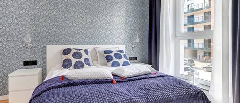 A bedroom with a patterned wallpaper, a large window with curtains, a bed with a blue blanket, and a white nightstand.