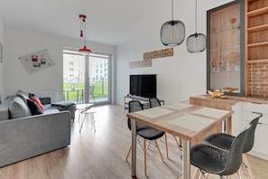 A modern living room with a grey sofa, a dining table with black chairs, a TV, and a large window with a view of a playground.