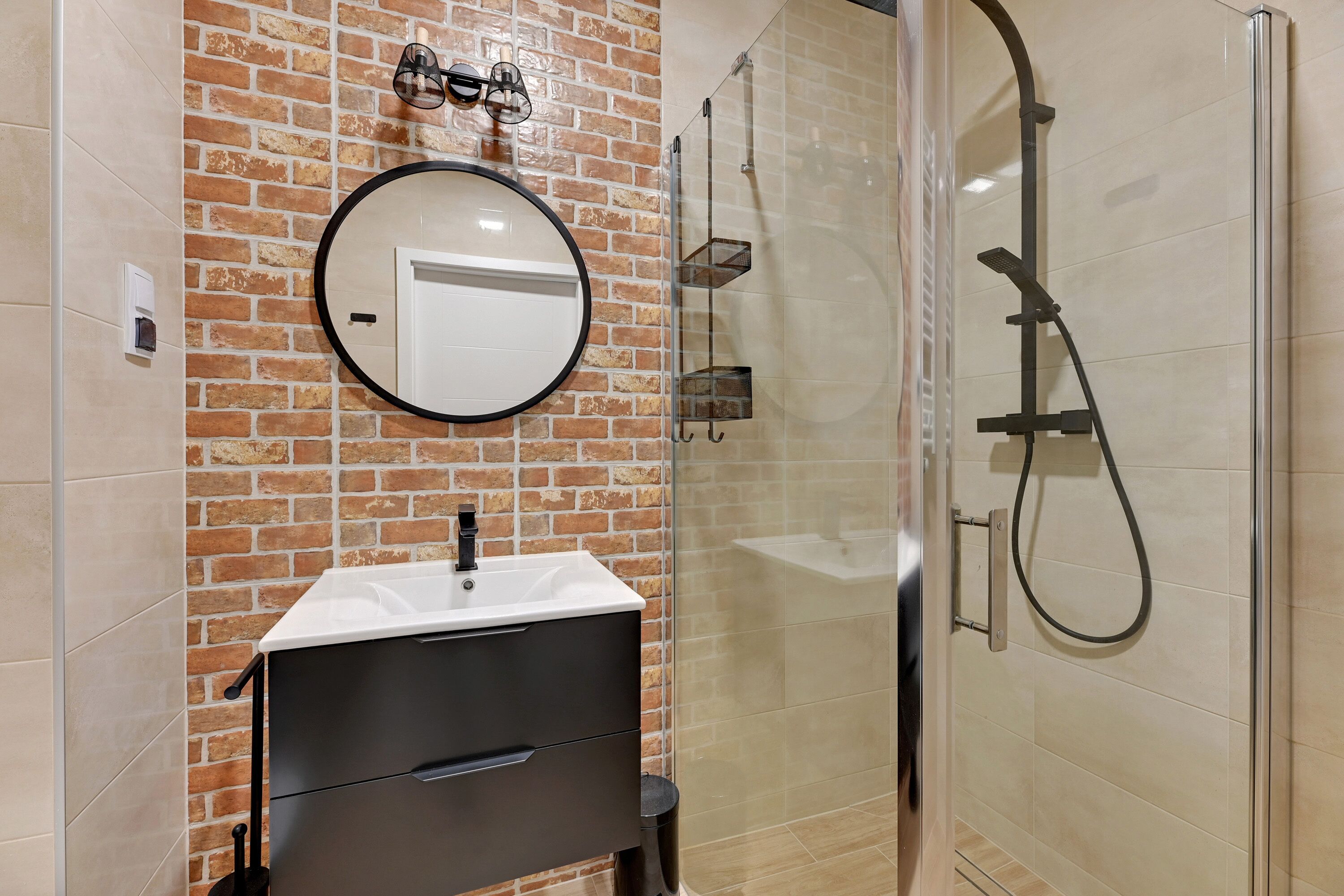 Un bagno con lavandino bianco, mobile contenitore nero e specchio rotondo. È presente un box doccia in vetro con soffione a mano.