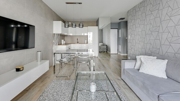 Una sala de estar moderna con un sofá gris, una mesa de centro de cristal transparente y un televisor de pantalla plana montado en la pared.