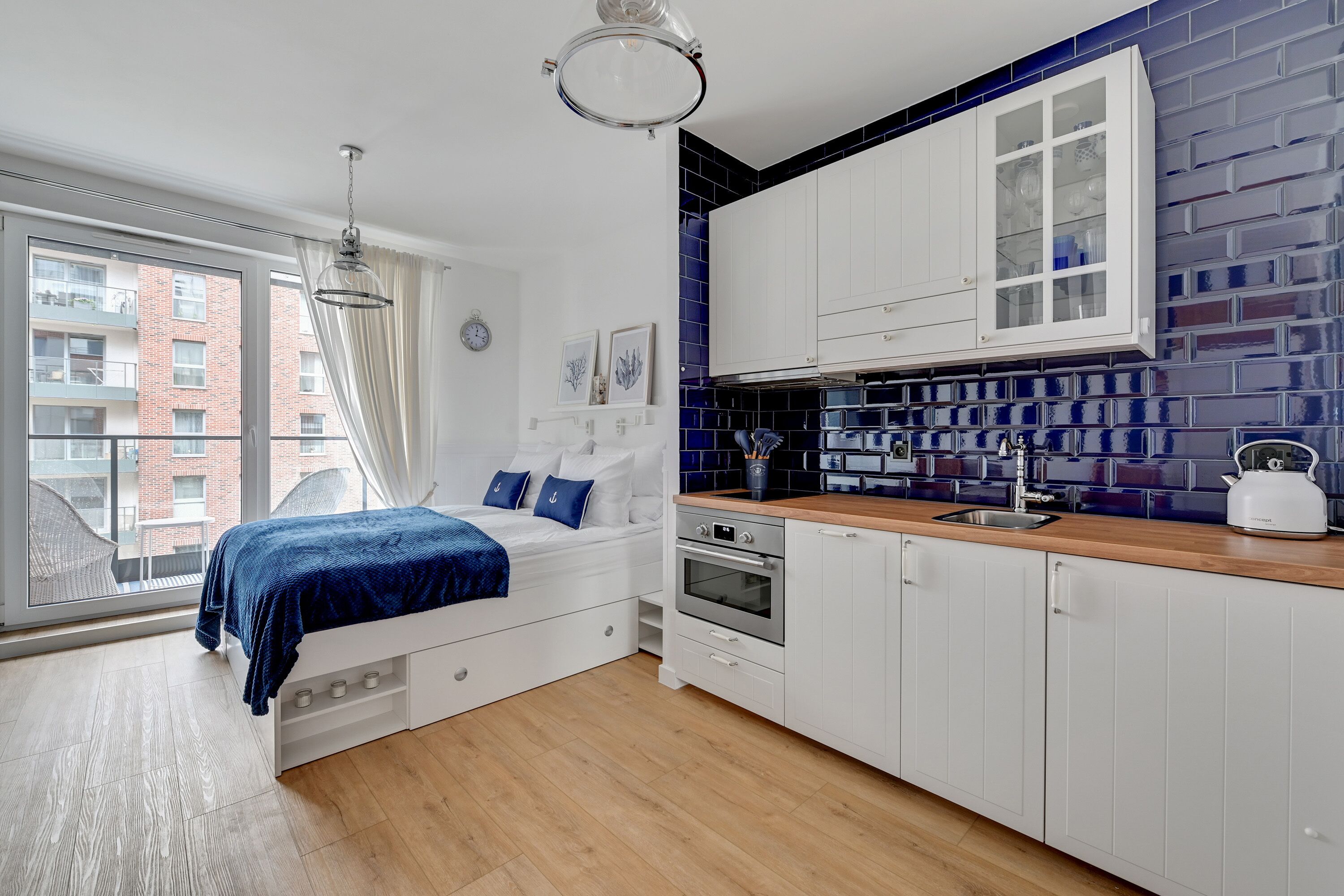 A modern bedroom with a large window, a bed with blue bedding, and a kitchenette with white cabinets and blue backsplash.