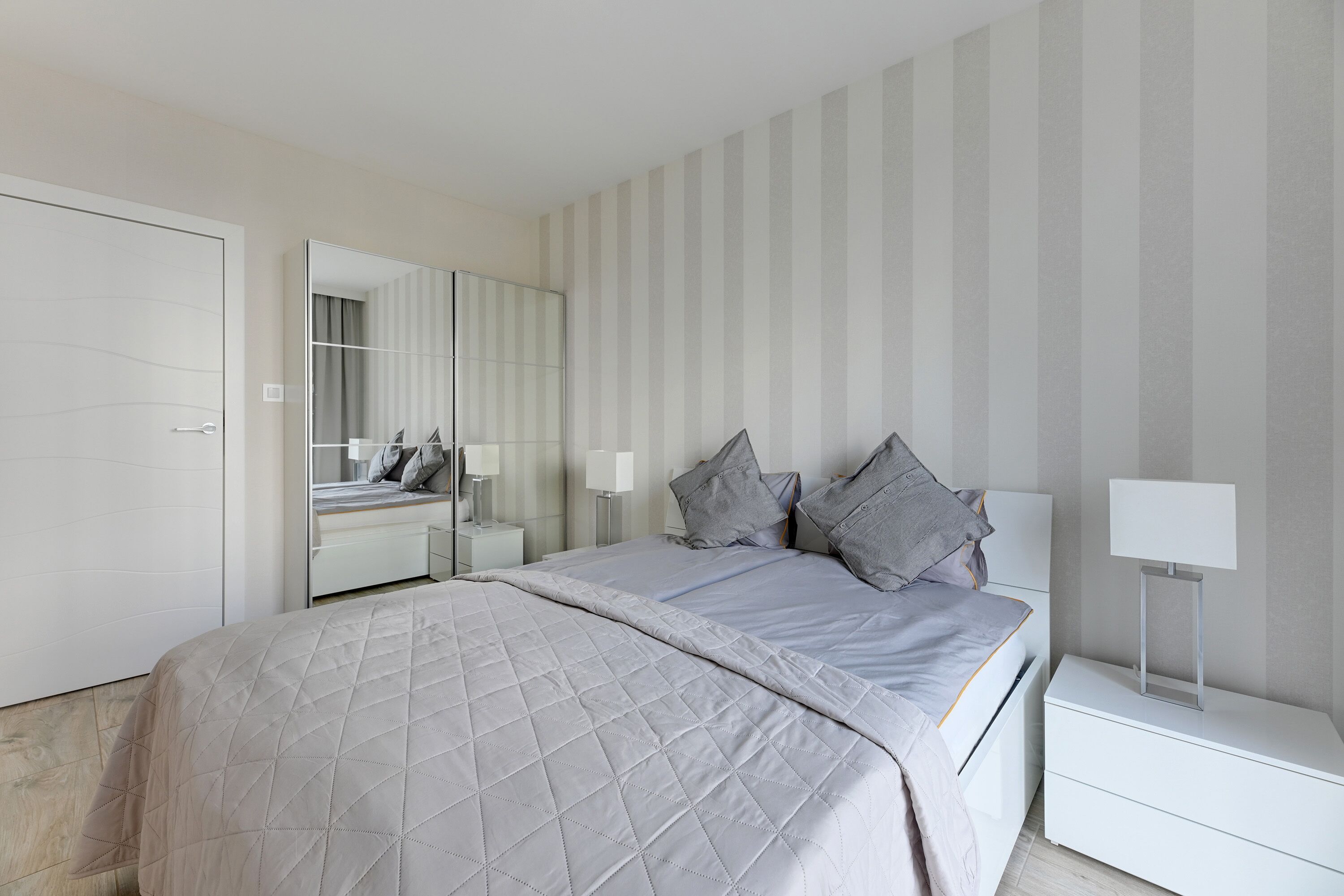A modern bedroom with a large bed, a nightstand, a bedside table, a mirror, and a striped wall.