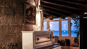 A rustic room with a piano, a framed picture on the wall, and a view of the sea through large windows. - Windmill Tower Beach House in Rhodes island (Ialisos)