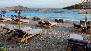 A beach with wooden lounge chairs and thatched umbrellas, arranged on a pebbled surface. - Windmill Tower Beach House in Rhodes island (Ialisos)