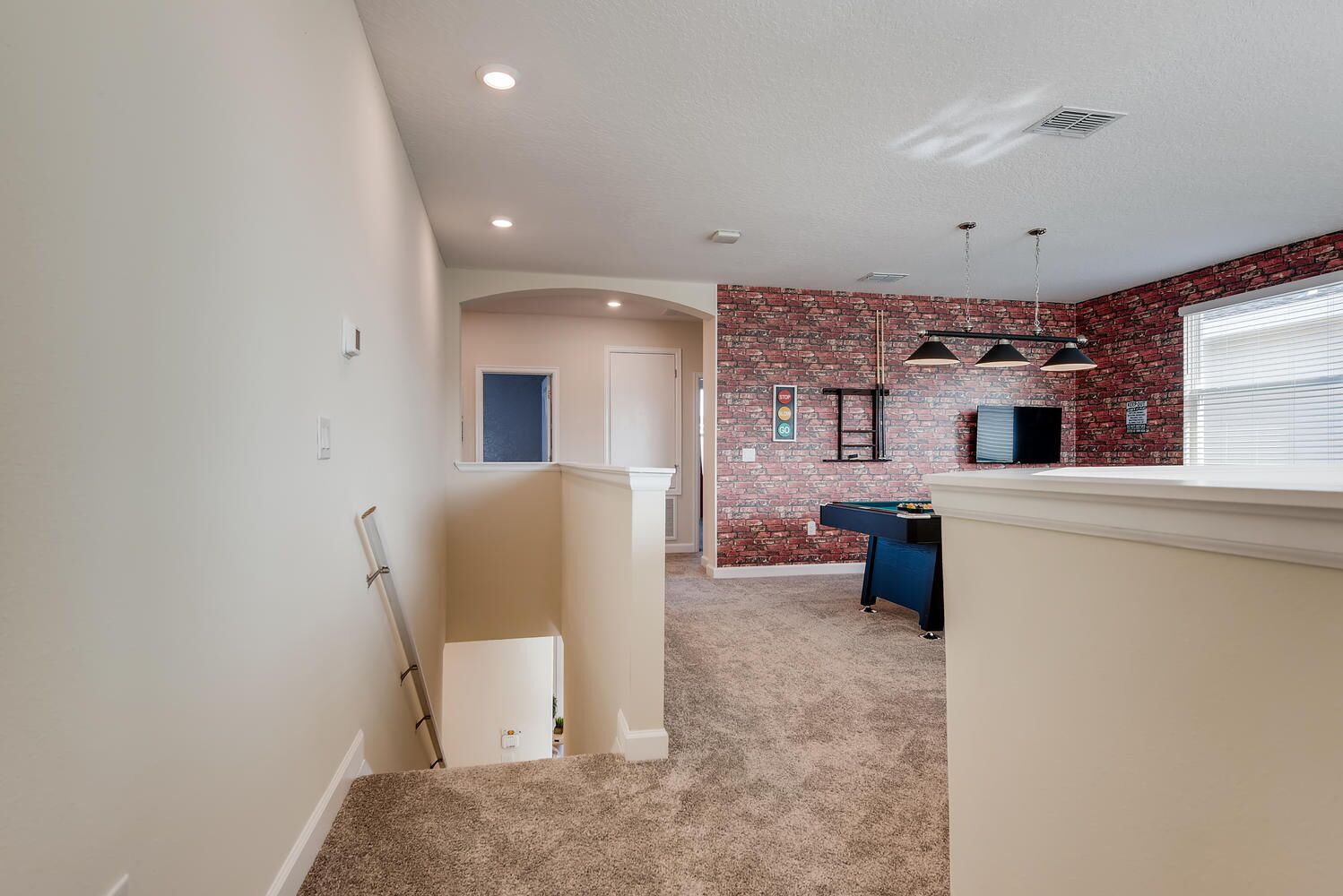 A hallway leading to a room with a pool table, a brick wall, and a window with blinds.
