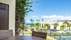A wooden table and chairs on a balcony with a view of a parking area, palm trees, and a building with a red roof.