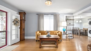A spacious living room with a wooden floor, a sofa, a coffee table, a dining area with a chandelier, and a laundry room with a washing machine.