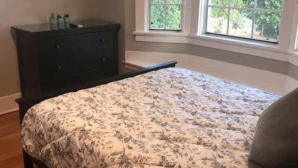 A bedroom with a bed, a dresser, a TV mounted on the wall, and a window with blinds.