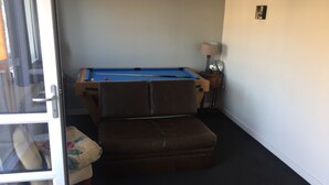Habitación con mesa de billar, un reposapiés de cuero marrón, una lámpara y una puerta que da a otra habitación.