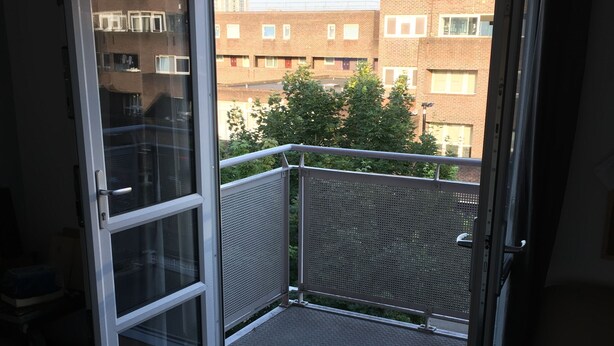 Una puerta corrediza de vidrio abierta que da a un balcón con barandilla de metal, con vista a un edificio de ladrillo y un árbol al fondo.