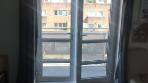 Una habitación con un gran ventanal corredero que da a un balcón exterior. La habitación tiene una alfombra oscura y una silla con un control remoto encima.