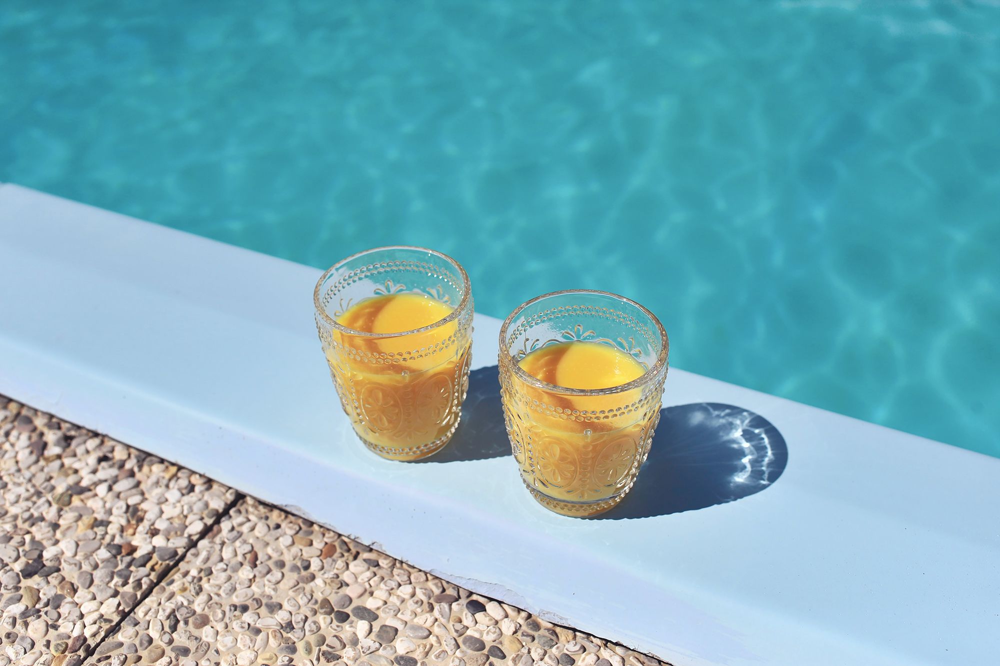 Two glasses of orange juice on a white ledge by a pool.