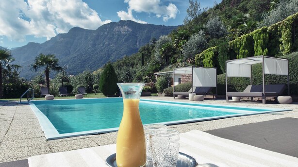 A poolside setting with a clear view of mountains, a lounge area, and a table with a drink and glass. - Das Wanda (Caldaro Sulla Strada del Vino)