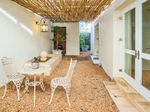 A covered outdoor seating area with a table and chairs, a decorative plant, and a stone pathway leading to a door.