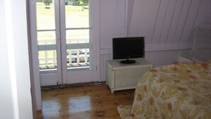Quarto com piso de madeira, uma televisão sobre uma mesinha branca e uma cama com colcha florida.