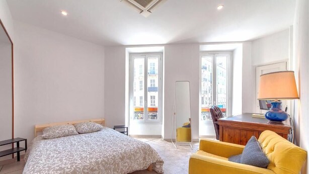 A bedroom with a bed, a yellow armchair, a desk with a lamp, and a view of buildings through large windows. - Perfect large beautiful apartment for Families with Children in Centre of Nice (Nice)