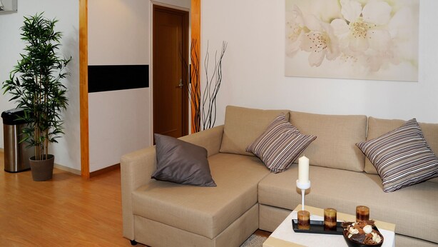 Ein Wohnzimmer mit einem beige farbenen Sofa, gestreiften Kissen, einer weißen Wand mit einem floralen Gemälde, einer Bambuspflanze und einem Tisch mit Kerzen und einer Schüssel.