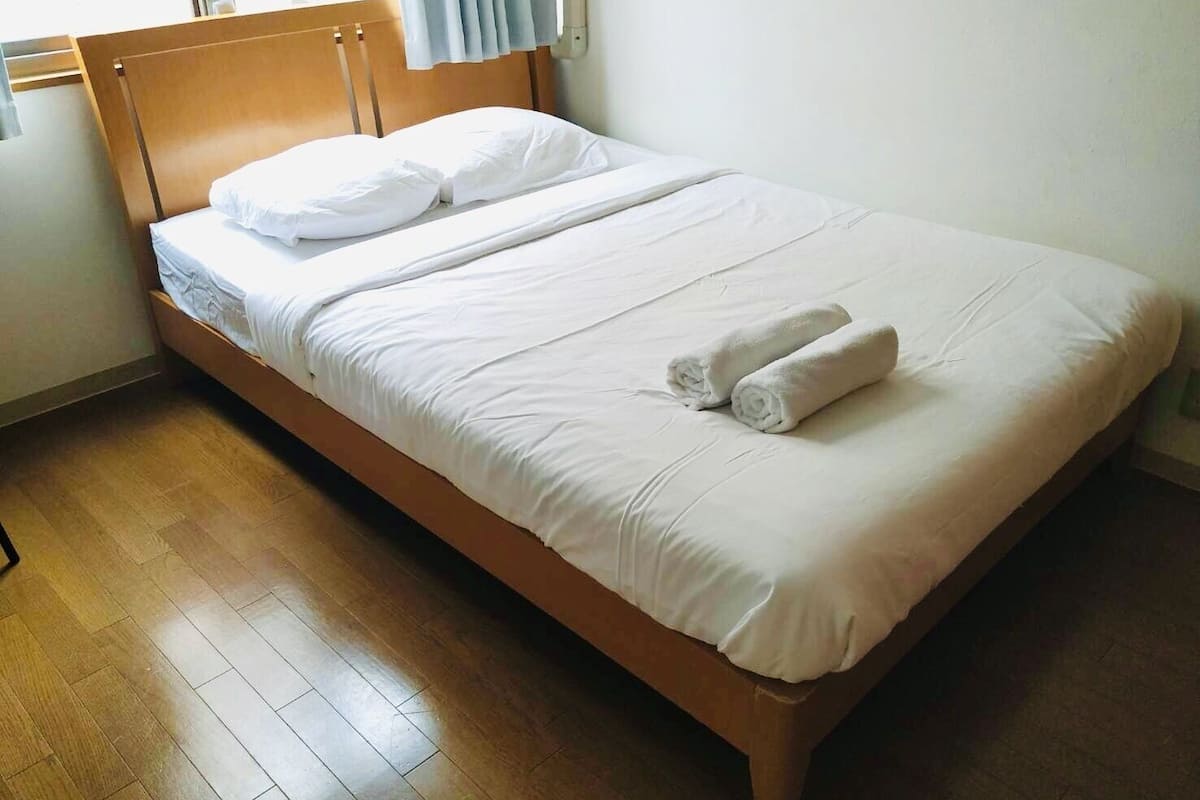 A neatly made bed with white linens and two rolled towels, a wooden headboard, and a wooden floor.