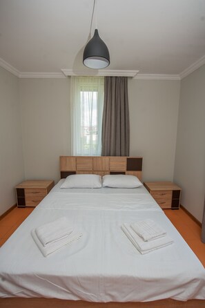 Cama bem arrumada com lençóis e toalhas brancos, cabeceira de madeira, mesas de cabeceira e um abajur suspenso.