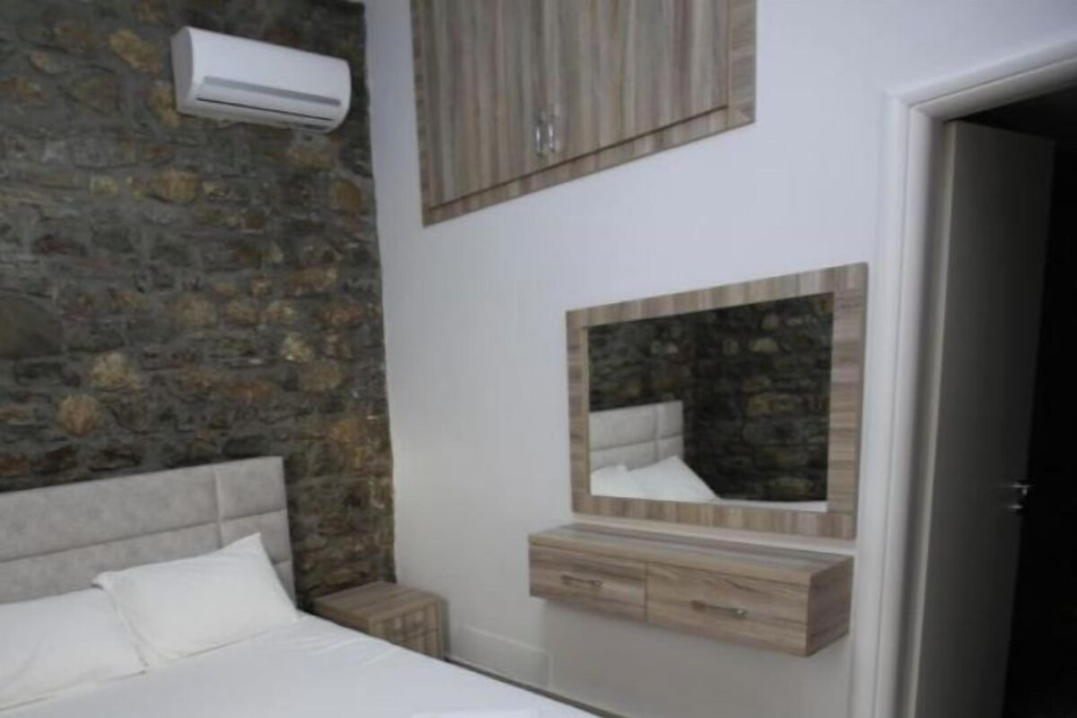 Un dormitorio con pared de piedra, cama con ropa de cama blanca, cabecero de madera, mesita de noche, cómoda y espejo.