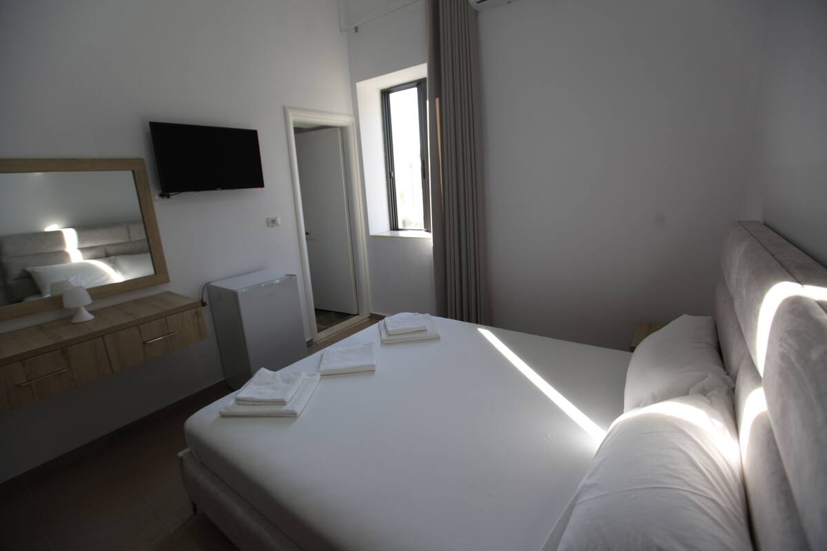 A hotel room with a bed, a TV mounted on the wall, a window with curtains, and a mirror above the dresser.