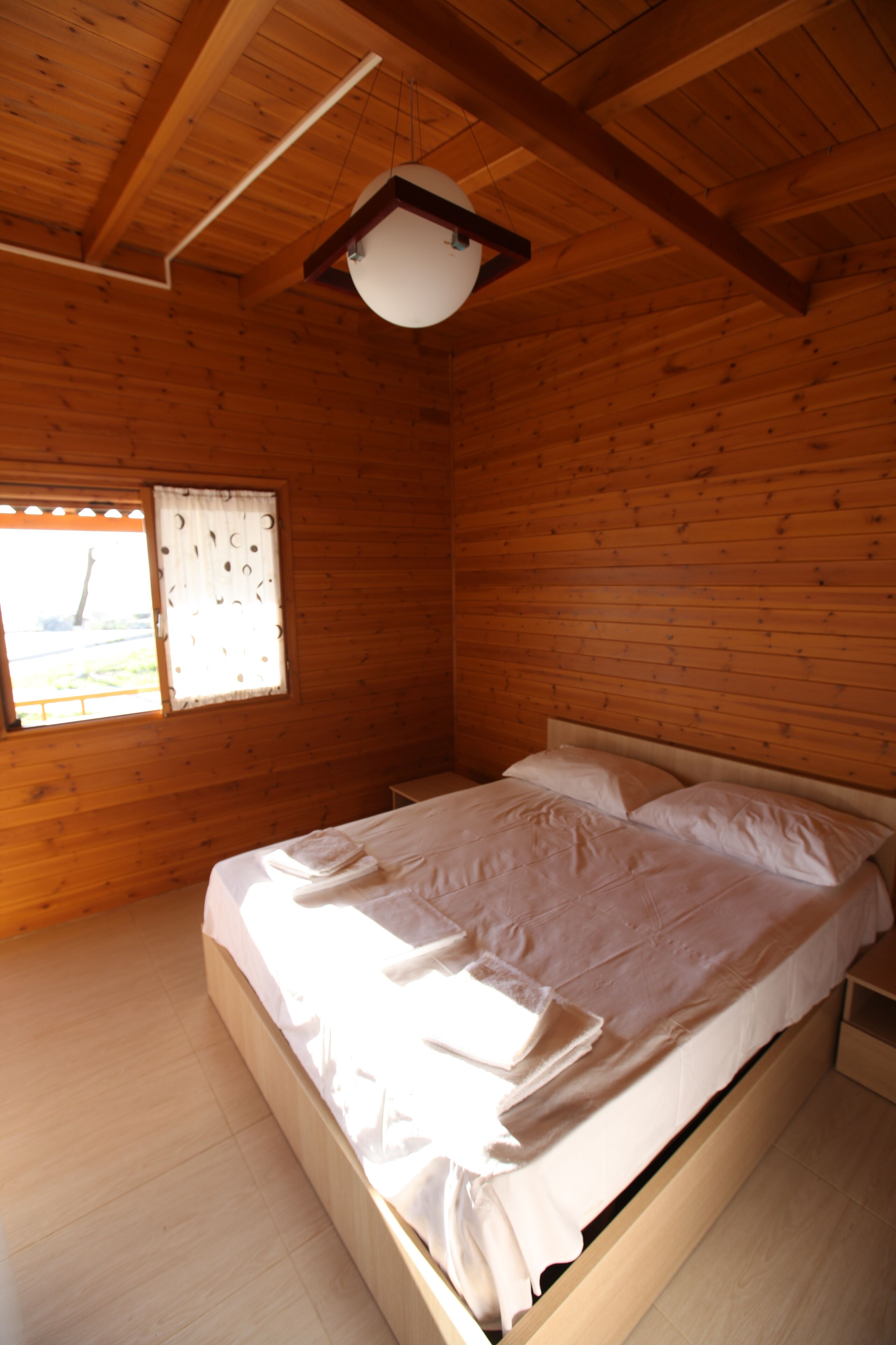 A wooden cabin bedroom with a single bed, a window with curtains, and a ceiling light fixture.