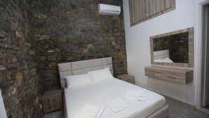 A bedroom with a stone wall, a bed with white linens, a wooden nightstand, a wooden dresser, and a mirror. - Bunec Beach Resort (Lukovë)