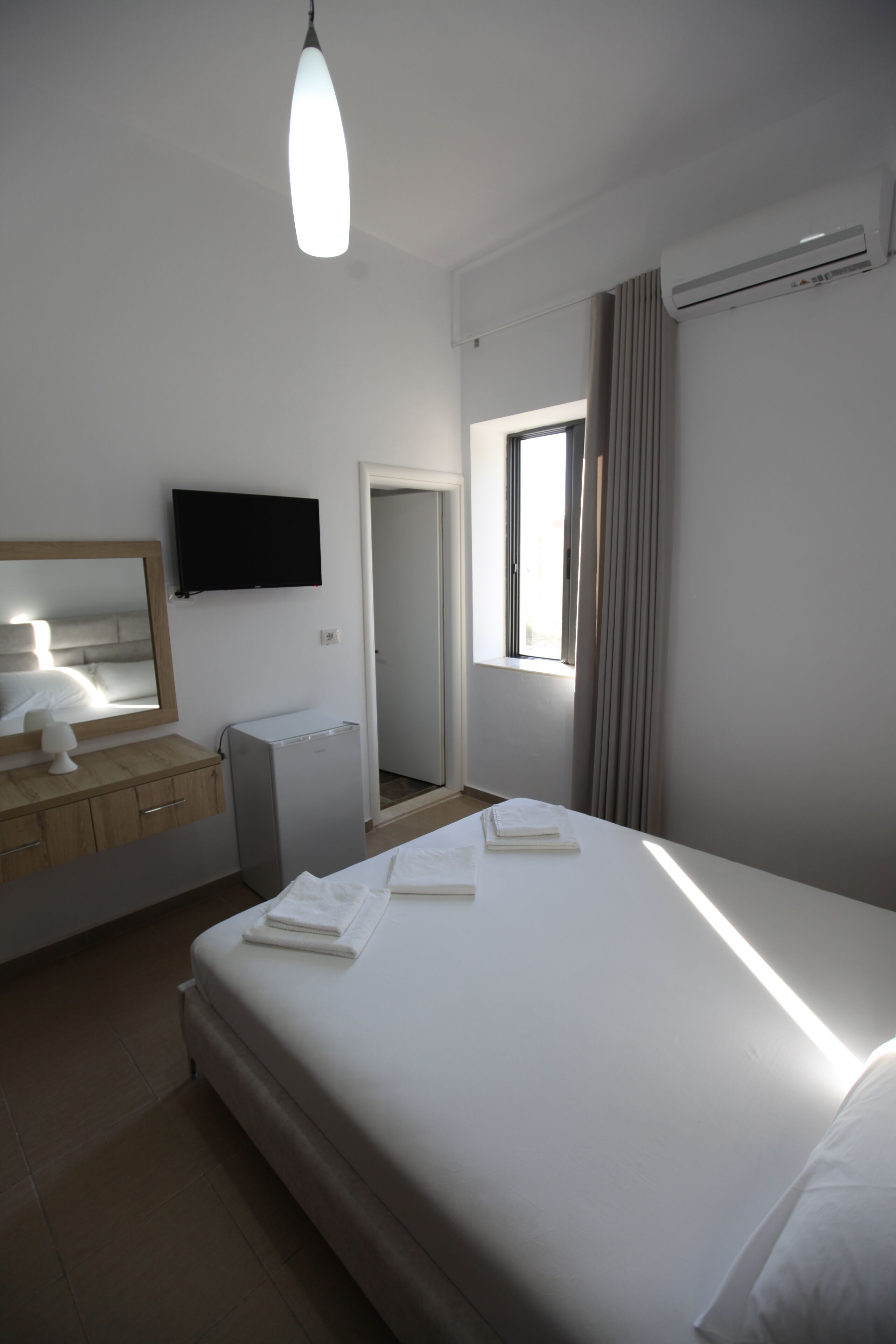 A modern hotel room with a large bed, a flat-screen TV mounted on the wall, a small desk with a lamp, and an air conditioning unit.