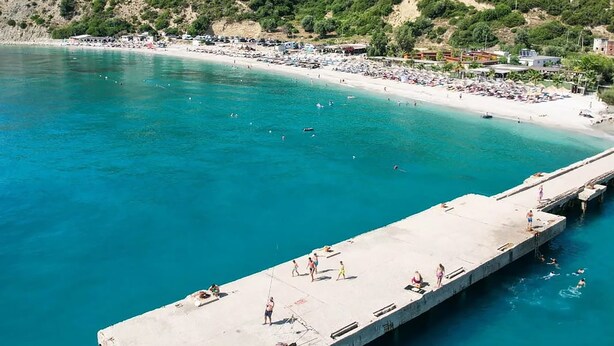 A coastal scene with a pier extending into turquoise waters, surrounded by a beach and hills in the background. - Bunec Beach Resort (Lukovë)