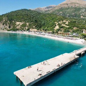 A coastal scene with a pier extending into turquoise waters, surrounded by a beach and hills in the background. - Bunec Beach Resort (Lukovë)