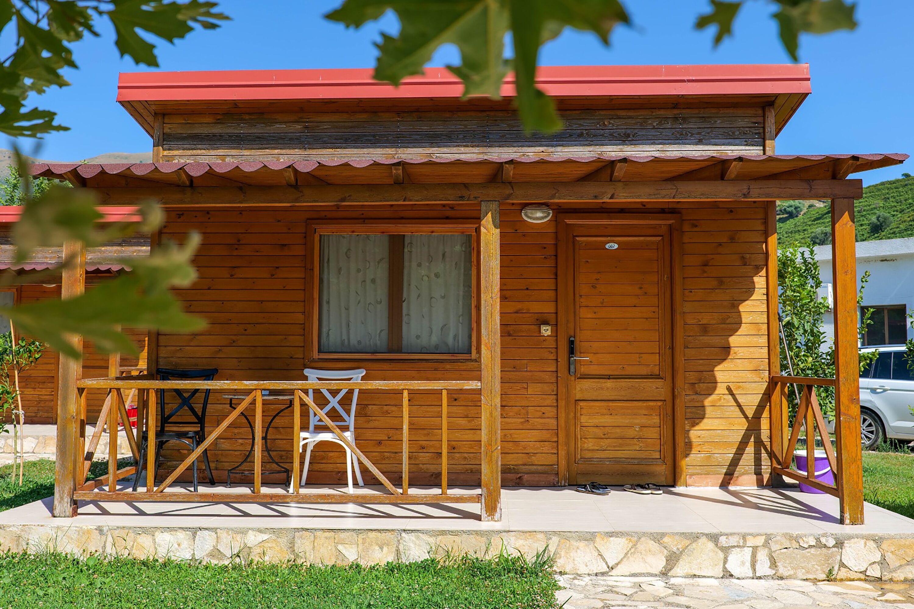 Una cabaña de madera con techo rojo, un porche con una silla blanca y cimientos de piedra.
