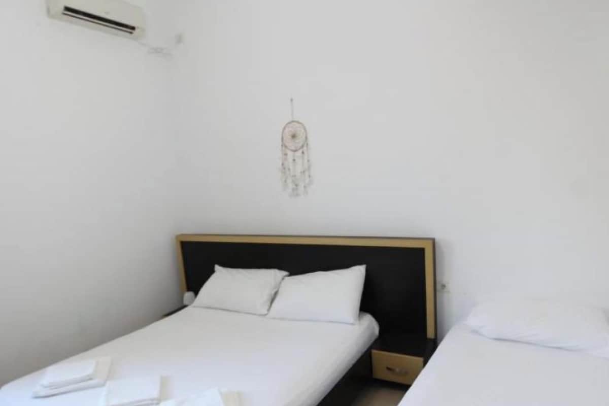 A room with two beds, white bedding, a wooden headboard, a wall-mounted air conditioner, and a dreamcatcher decoration.