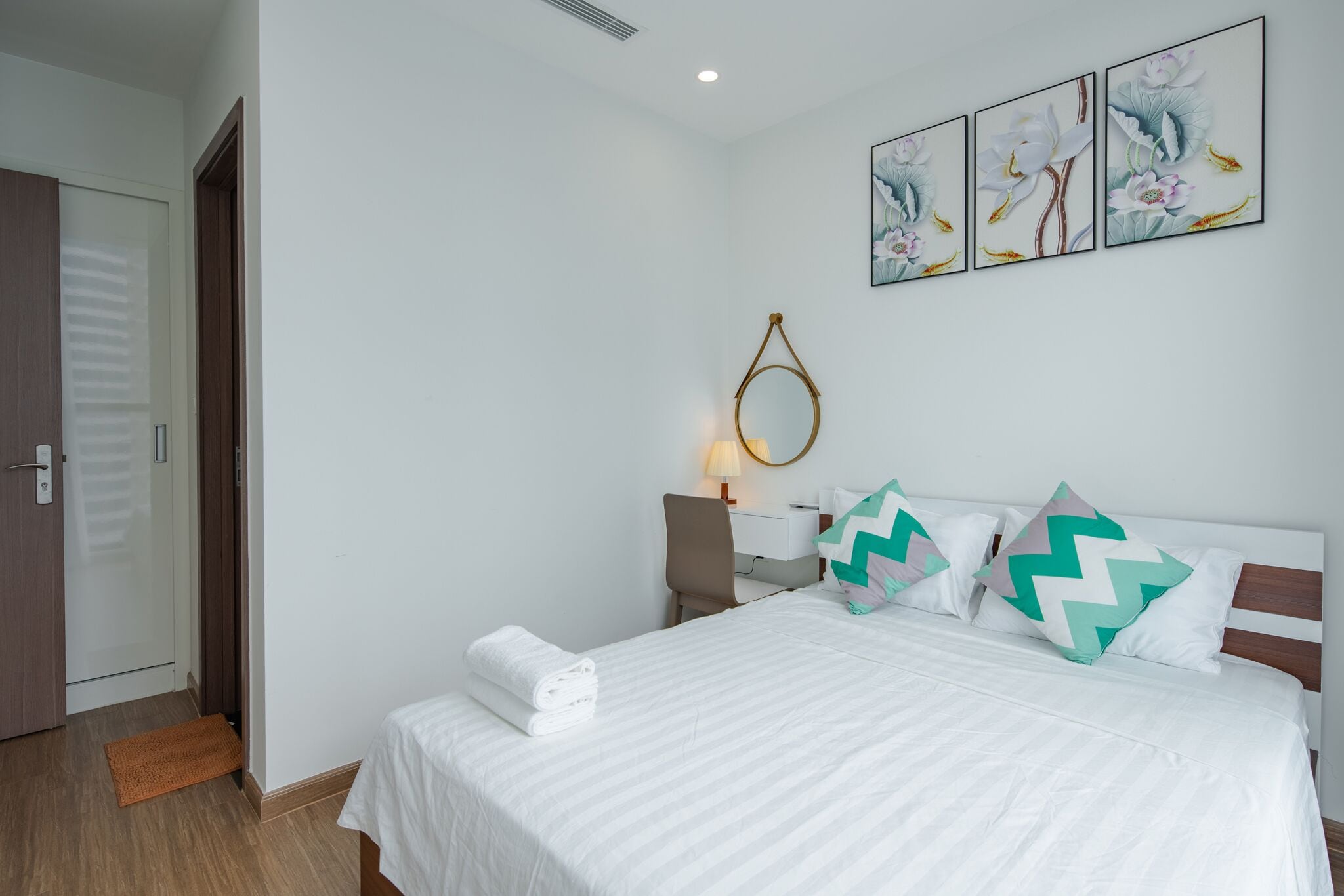 A neatly made bed with white bedding and decorative pillows, a bedside table with a lamp, and a wall-mounted mirror.