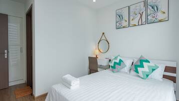 A neatly made bed with white bedding and decorative pillows, a bedside table with a lamp, and a wall-mounted mirror.