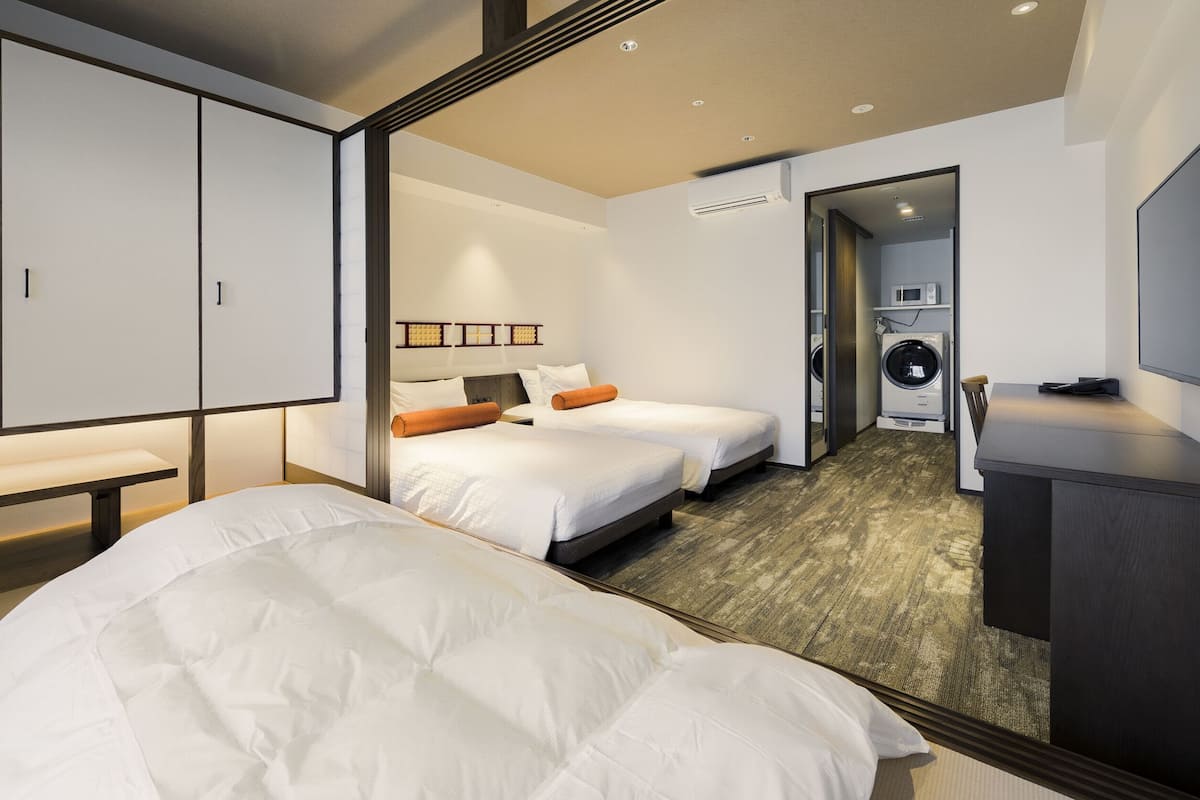 A hotel room with two beds, a bench, a TV, and a washing machine in the background.