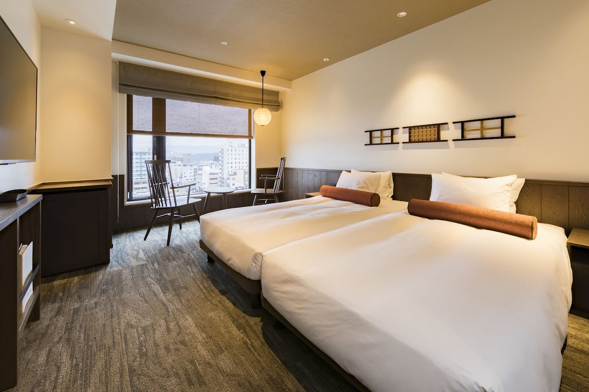A hotel room with two beds, a desk, a chair, a TV, and a large window with a city view.
