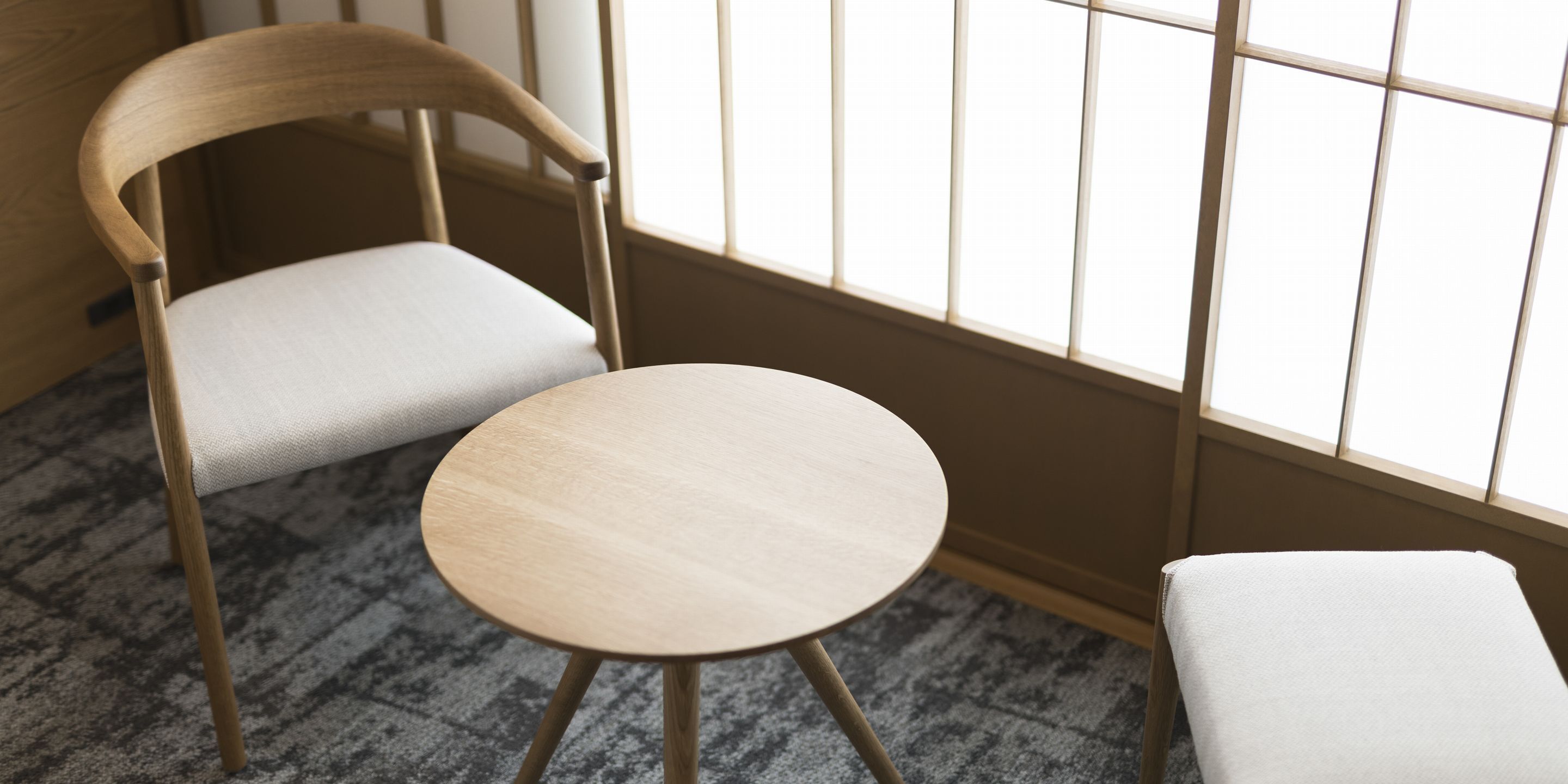 A chair with a cushioned seat and back, a small round table, and a matching ottoman, all in a room with a patterned carpet and a window with vertical blinds.