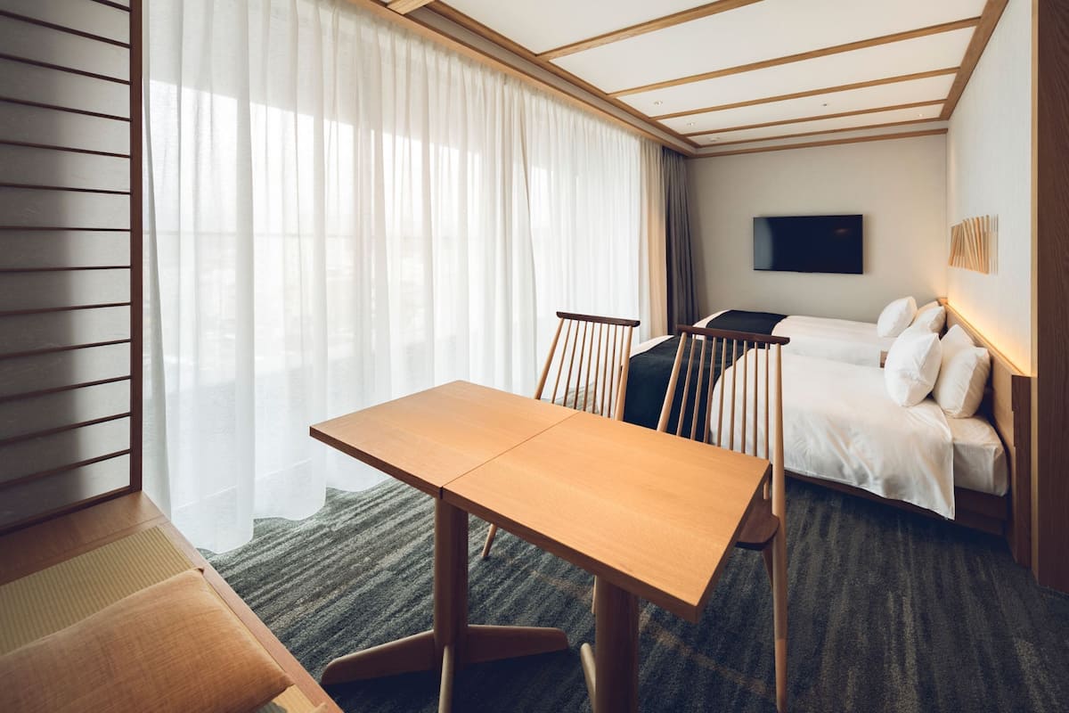 A hotel room with a wooden table, a bed with white bedding, a TV mounted on the wall, and sheer curtains on the window.