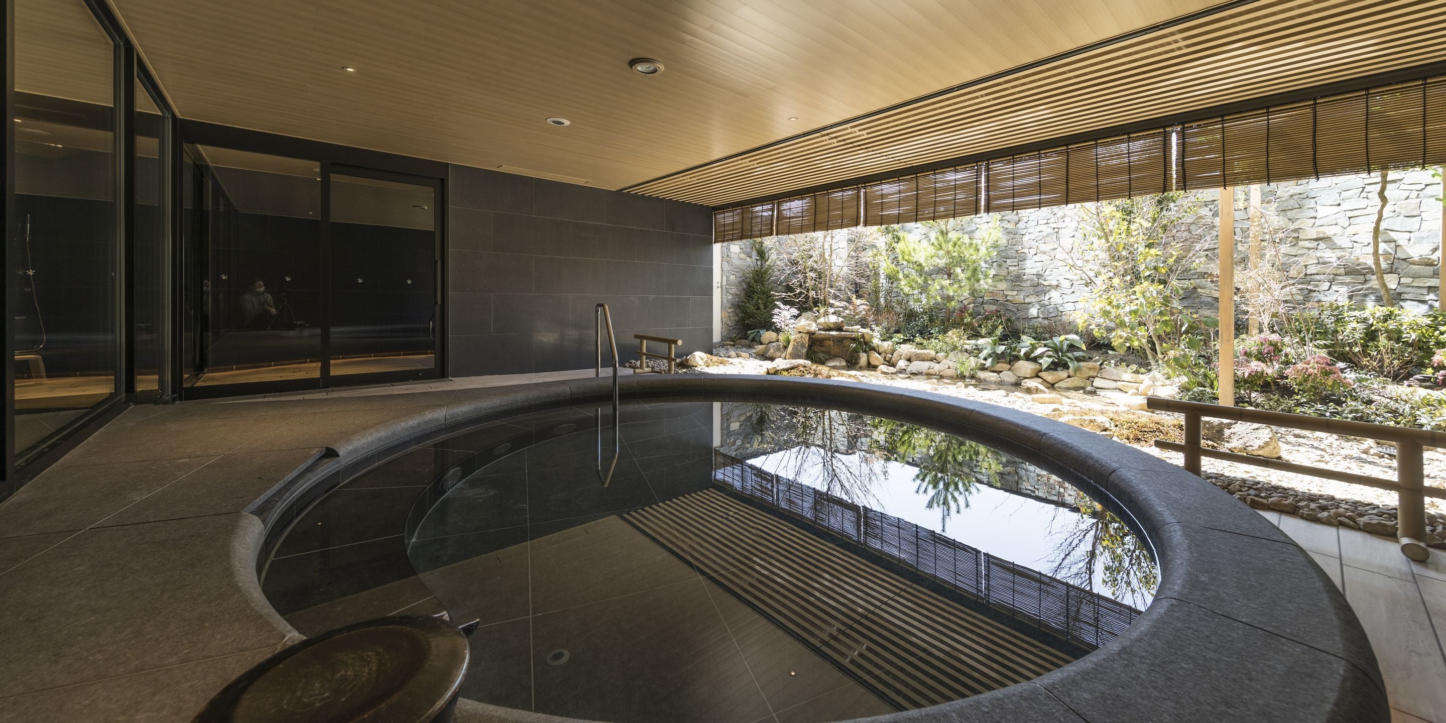 A modern indoor swimming pool with a curved design, surrounded by a stone wall and a wooden deck.