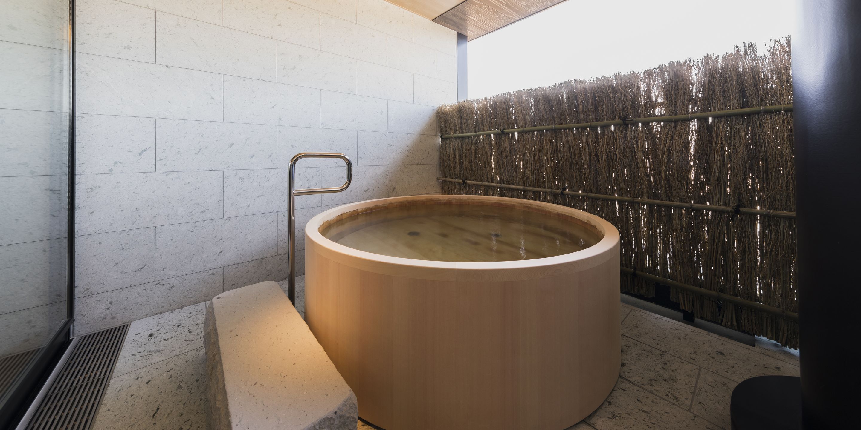 A wooden hot tub in a spa area with a tiled wall and a bamboo fence.